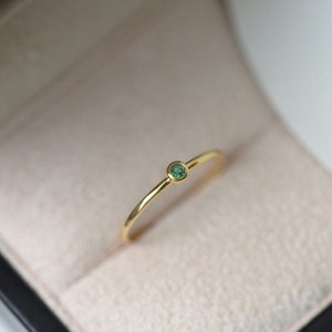 Può includere: Un delicato anello d'oro con una piccola gemma rotonda verde smeraldo. L'anello ha un design minimalista ed è presentato in un portagioie con interno beige chiaro. Il gambo dell'anello è sottile e lucido.