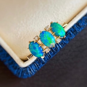 Könnte beinhalten: Ein goldener Ring mit drei ovalen Opalen und kleinen Diamanten, die in die Ringschiene eingefasst sind. Die Opale haben eine leuchtende blaugrüne Farbe.