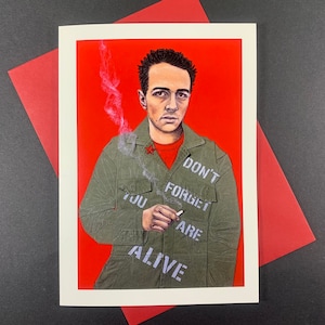 May include: A man with short dark hair is wearing a green jacket with the words "Don't Forget You Are Alive" written on it. He is holding a cigarette in his right hand and has a red star pin on his jacket. The background is red.