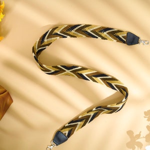 May include: A woven bag strap with a chevron pattern in shades of olive green, brown, black, and white. The strap has black leather ends with silver-colored metal clasps. The strap is shown on a beige background.