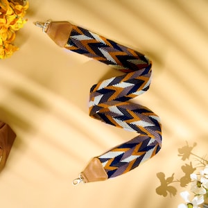May include: A woven bag strap featuring a repeating chevron pattern in navy blue, mustard yellow, grey, and white. The strap includes tan faux leather end tabs and silver-toned metal swivel lobster clasps at both ends for secure attachment.