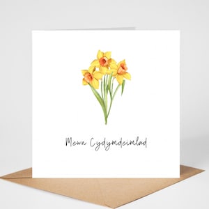 May include: A white greeting card featuring a watercolor illustration of yellow daffodils with orange centers and green stems. The card has the text "Mewn Cydymdeimlad" in black script. The card is on a brown envelope.