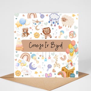 May include: A greeting card with a baby theme. The card features illustrations of a teddy bear, lion, sheep, rainbow, and balloons. The text "Croeso Ir Byd" is written on a brown banner. The card is on a brown envelope.