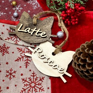 May include: Two wooden cat-shaped ornaments with the names "Latte" and "Roscoe" written on them. The ornaments are on a red and white fabric background with pine cones and red berries.