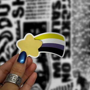 May include: A sticker in the shape of a shooting star with a yellow star and a rainbow tail in the colors of the non-binary pride flag: yellow, white, purple, and black. The sticker is held in front of a blurred black and white background.