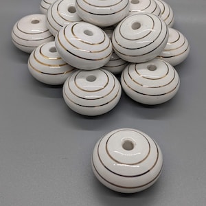May include: A pile of white ceramic beads with gold stripes. The beads have a hole in the center and are approximately 1 inch in diameter.