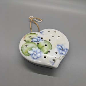 Könnte beinhalten: Weißer Keramik-Anhänger in Herzform mit einem floralen Design aus blauen und grünen Veilchen. Der Anhänger hat eine goldene Schnur zum Aufhängen und Löcher am Rand.