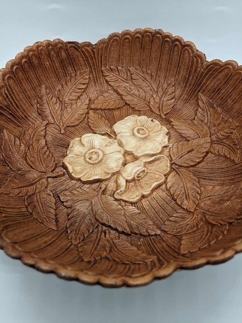 Puede incluir: Un cuenco de madera marr&oacute;n con un dise&ntilde;o tallado de hojas y flores. Las flores son blancas y tienen una textura en relieve.