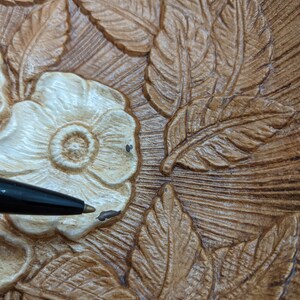 Puede incluir: Primer plano de un plato decorativo de madera con un dise&ntilde;o floral tallado. El plato presenta una flor blanca con un centro marr&oacute;n y hojas de color marr&oacute;n y beige.
