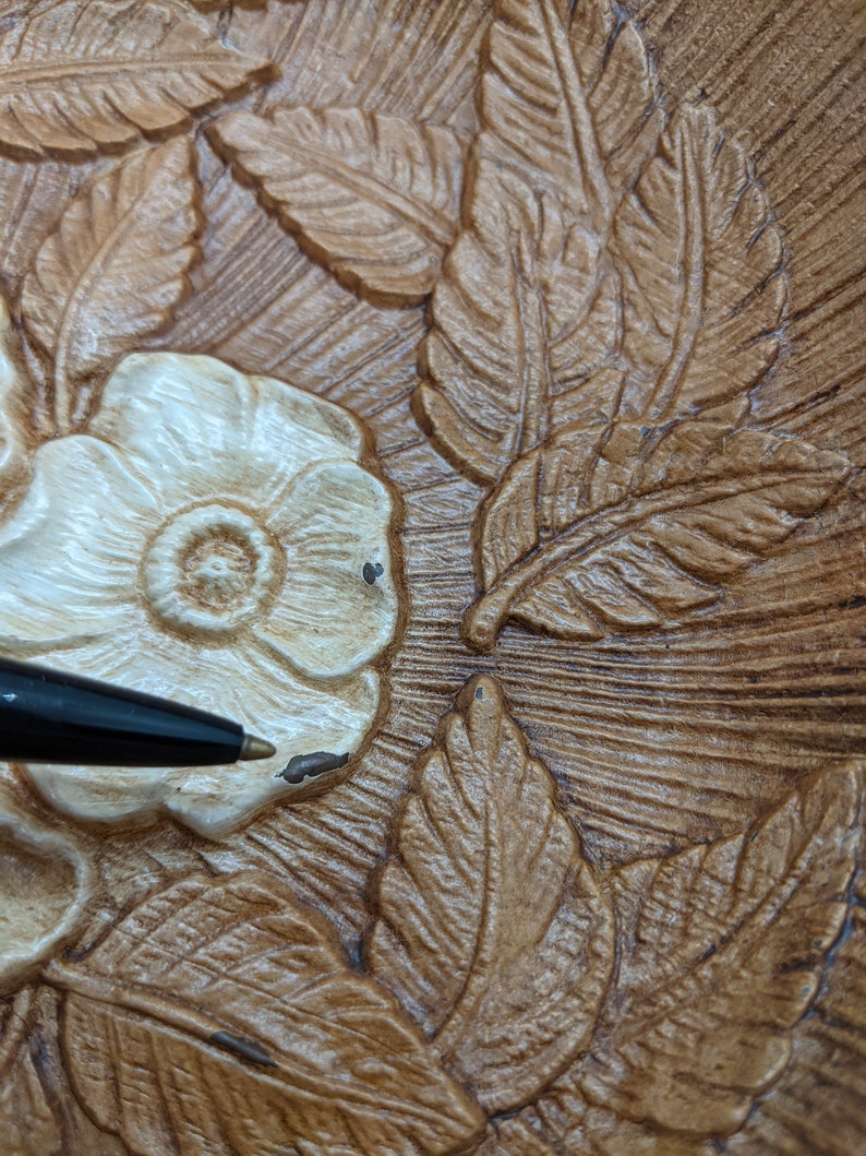 Puede incluir: Primer plano de un plato decorativo de madera con un dise&ntilde;o floral tallado. El plato presenta una flor blanca con un centro marr&oacute;n y hojas de color marr&oacute;n y beige.