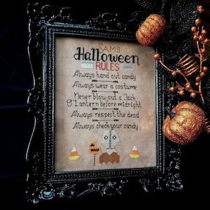 May include: A framed cross-stitch design with a black frame and a beige background. The design features a stitched image of a character from the movie Halloween, holding a jack-o-lantern. The text reads "Sam's Halloween Rules" and includes a list of rules for Halloween, such as "Always hand out candy" and "Always wear a costume".
