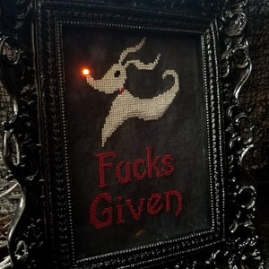 Puede incluir: Un bordado de punto de cruz enmarcado en negro con una imagen bordada en blanco de Jack Skellington de la película Pesadilla antes de Navidad. El texto "F**ks Given" está bordado en rojo debajo de la imagen.