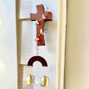 May include: A wooden cross with a decorative design, featuring a dark brown finish and pink leaf accents. Below the cross hangs a dark brown semi-circle with three hanging elements: two gold-coloured bells and a white, translucent gem.