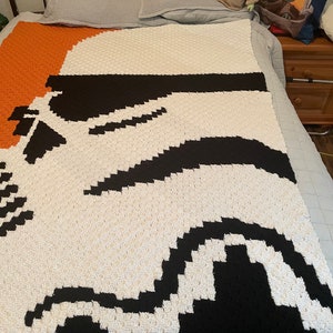 May include: A white, black, and orange crocheted blanket featuring a Star Wars Stormtrooper design.