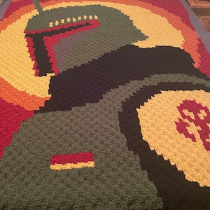Puede incluir: Una manta de ganchillo con una imagen pixelada de Boba Fett de Star Wars. La manta está hecha con una variedad de colores, incluyendo verde, marrón, amarillo, naranja y rojo.