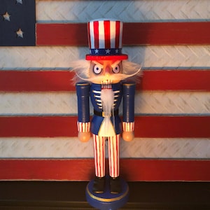 May include: A wooden nutcracker figurine dressed as Uncle Sam. The nutcracker has a red, white, and blue hat, a blue jacket, and white pants with red stripes. The nutcracker has a white beard and mustache, and a scary expression on its face.
