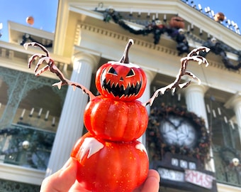 May include: A three-tiered pumpkin snowman decoration with a jack-o'-lantern face on the top pumpkin. The snowman is holding out its arms, which are made of branches with white frost.