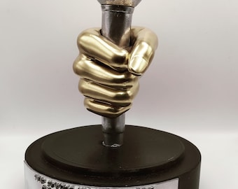 May include: A trophy shaped like a microphone, featuring a silver body and a gold-coloured hand grasping the shaft. The microphone's top has a silver sphere with a gold "S" emblem. The black base has silver text: "THE SOURCE HIP-HOP AWARDS PRODUCER OF THE YEAR".
