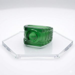 Könnte beinhalten: Ein leuchtend grüner Glasring mit zylindrischem Design, einem kreisförmigen Mittelteil und rechteckigen Akzenten. Der Ring wird auf einem transparenten, sechseckigen Acrylständer vor weißem Hintergrund präsentiert. Das Design des Rings erinnert an eine Laterne.