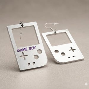 May include: Silver-colored earrings in the shape of a Game Boy. The earrings have a rectangular shape with a screen cutout and the words "GAME BOY" in purple. The design includes buttons and a cross-shaped control.