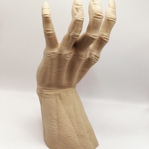 May include: A beige, stylized hand sculpture with long, slender fingers. The hand is posed in a gesture, with the fingers slightly curved. The sculpture has a textured surface, and is set against a white background.