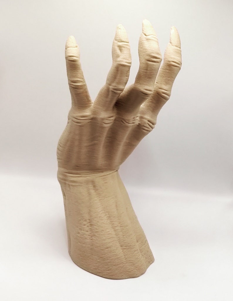 May include: A beige, stylized hand sculpture with long, slender fingers. The hand is posed in a gesture, with the fingers slightly curved. The sculpture has a textured surface and is set against a white background.