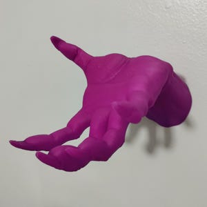 May include: A magenta-colored hand sculpture, with fingers extended, mounted on a white wall. The hand is detailed, showing the texture and form of a human hand. The sculpture is a unique decorative item.
