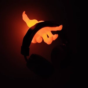 May include: Glowing orange hand-shaped light fixture, seemingly emerging from a black headphone. The light emits a warm, vibrant glow against a dark background, highlighting the hand's form and the headphone's silhouette.