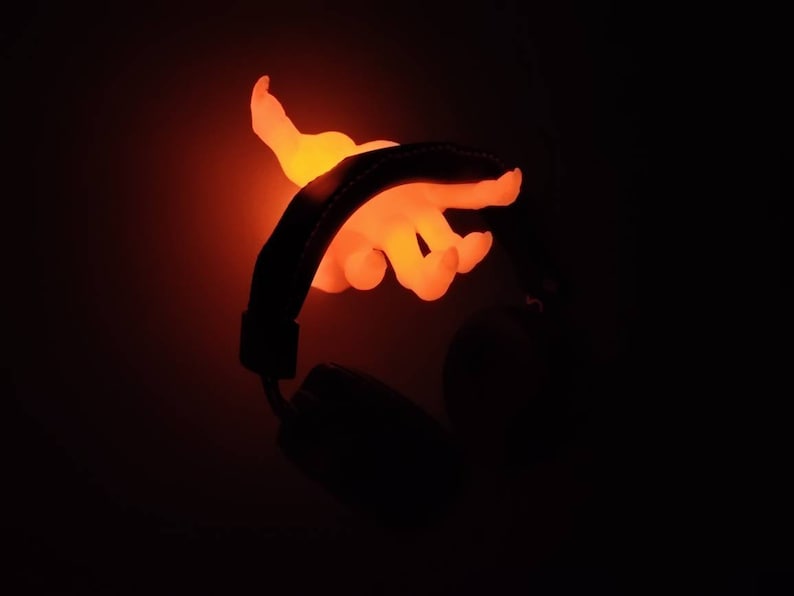 May include: Glowing orange hand-shaped light fixture, seemingly emerging from a black headphone. The light emits a warm, vibrant glow against a dark background, highlighting the hand's form and the headphone's silhouette.