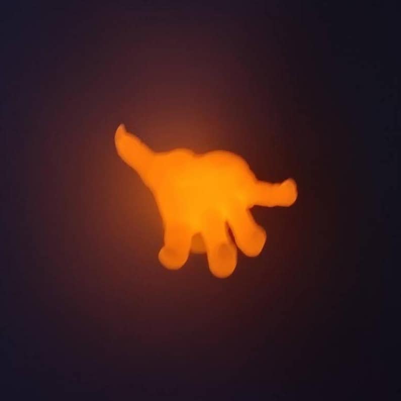 May include: A glowing orange hand-shaped night light against a dark background. The light emits a warm, inviting glow, with the fingers and palm clearly defined. The overall effect is a soft, ambient illumination.