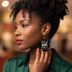 May include: A woman wearing black earrings with the words "Black Girl Magic" in white script. The earrings are in the shape of a woman's silhouette. The woman is wearing a green shirt.