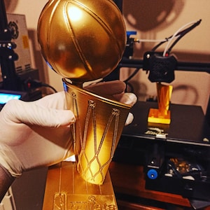May include: A gold-colored trophy with a basketball on top and a net-like design on the sides. The trophy has a rectangular base with the text "NBA Bullets". The trophy is held by a gloved hand.