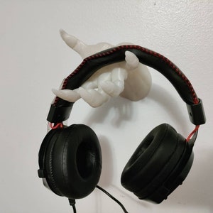 May include: Black over-ear headphones with red accents hanging from a white hand-shaped wall mount. The headphones have a padded headband with red stitching and large earcups. The wall mount is a sculpted hand.