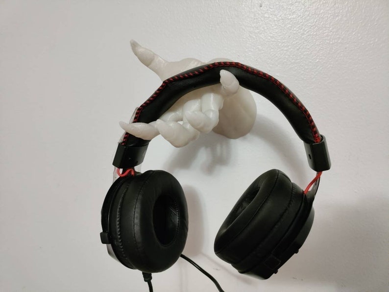 May include: Black over-ear headphones with red accents hanging from a white hand-shaped wall mount. The headphones have a padded headband with red stitching and large earcups. The wall mount is a sculpted hand.