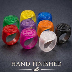 May include: A collection of nine translucent rings in various colors including red, green, yellow, purple, orange, blue, and white, plus one solid black ring. Each ring features a unique embossed symbol on the top face. The text HAND FINISHED is visible.