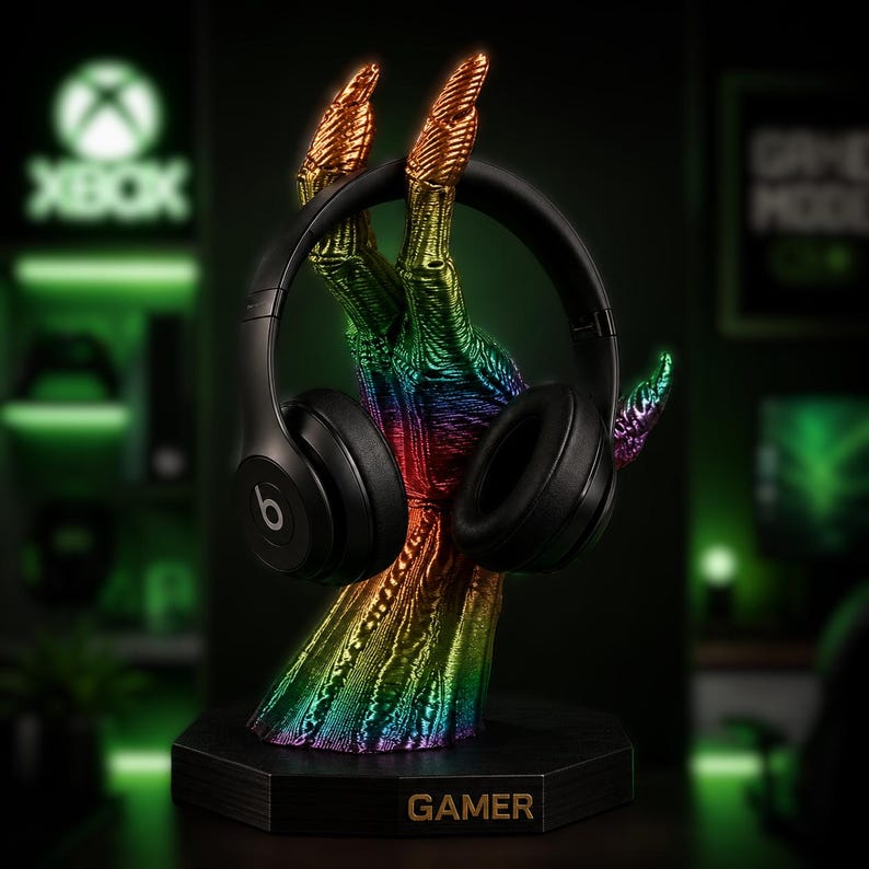 May include: A black Beats headphones rest on a rainbow-colored hand-shaped stand. The stand has a black hexagonal base with the word "GAMER" in gold lettering. The hand is detailed with gold fingertips.