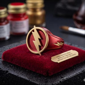 May include: A red and gold Flash ring with a lightning bolt emblem, displayed on a red velvet surface. The ring is part of the Artisan Collector Series. The ring is hand-painted resin.