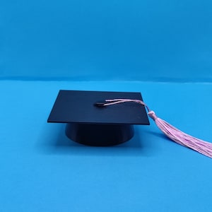 May include: A black graduation cap with a square top and a round base. A pink tassel hangs from the cap. The cap is set against a solid blue background.