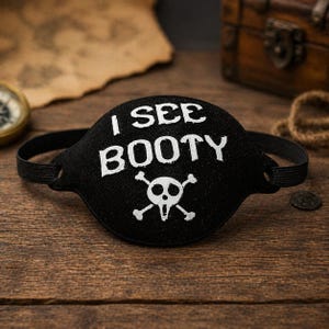 May include: Black pirate eye patch with the text "I SEE BOOTY" in white, along with a skull and crossbones graphic. The eye patch has a black elastic band. The background includes a treasure chest and a compass.