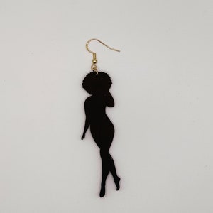 May include: A black silhouette earring featuring a woman with an afro hairstyle. The earring has a gold-coloured hook. The woman's silhouette is in a graceful pose, with one arm raised. The background is plain white.