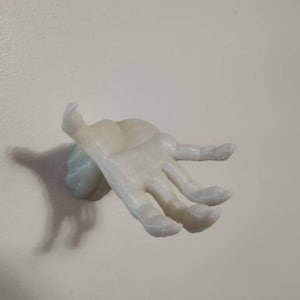May include: A light-colored, hand-shaped wall hook. The hook is mounted on a small, round base and extends outward with fingers slightly curved. The object is made of a smooth material and is mounted on a white wall.