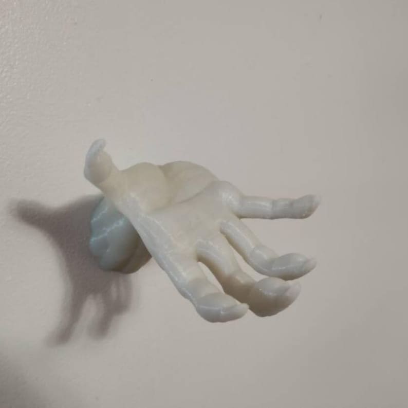May include: A light-colored, hand-shaped wall hook. The hook is mounted on a small, round base and extends outward with fingers slightly curved. The object is made of a smooth material and is mounted on a white wall.