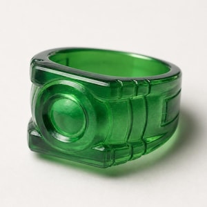 Op de afbeelding: Een opvallende groene ring met een geometrisch ontwerp. De ring is gemaakt van een doorschijnend materiaal, met een gedetailleerd, gebeeldhouwd uiterlijk. Het ontwerp van de ring heeft een cirkelvormig embleem en rechthoekige accenten.