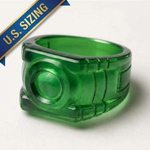 May include: A translucent green ring featuring a circular centre emblem and geometric side detailing. A blue banner in the upper left corner contains the text U.S. SIZING in yellow capital letters, indicating the ring follows American sizing standards.