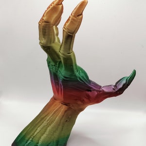May include: A 3D-printed hand sculpture with long, sharp fingernails. The hand is posed with two fingers extended upwards. The sculpture features a gradient of colors, transitioning from purple to green to gold. The hand is set against a white background.