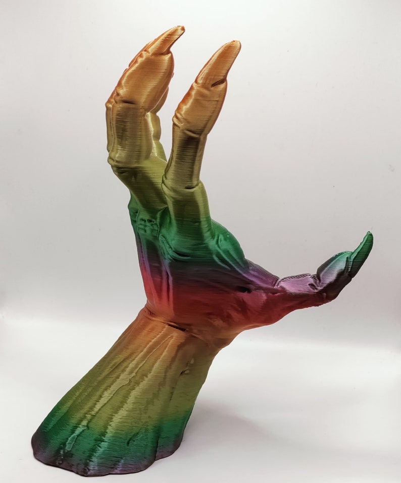 May include: A 3D-printed hand sculpture with long, sharp fingernails. The hand is posed with two fingers extended upwards. The sculpture features a gradient of colors, transitioning from purple to green to gold. The hand is set against a white background.