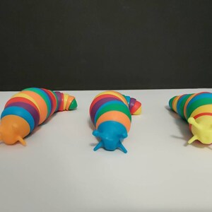 May include: Three colorful toy slugs with rainbow-striped bodies and contrasting heads. The slugs are arranged on a white surface against a dark background. Each slug has a different head color: orange, blue, and yellow.