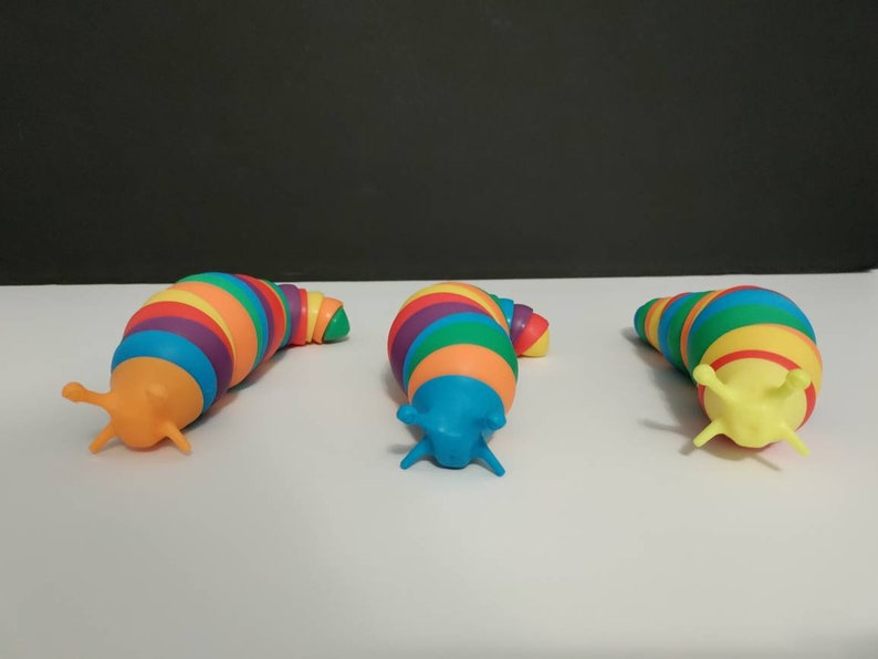 May include: Three colorful toy slugs with rainbow-striped bodies and contrasting heads. The slugs are arranged on a white surface against a dark background. Each slug has a different head color: orange, blue, and yellow.