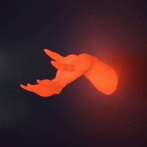 May include: An illuminated orange hand sculpture, mounted on a dark wall. The hand is posed as if reaching out, with fingers slightly curved. The lighting creates a warm, glowing effect, highlighting the form and texture of the hand.