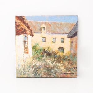 May include: Oil painting on canvas depicting a European village scene. The artwork features a building with a light-colored facade, a red-brown roof, and a blue sky. Bushes and flowers are in the foreground.